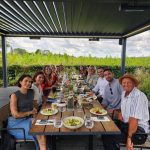 People sat around a table and underneath a louvered roof pergola