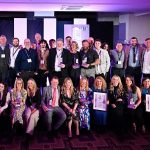 A large group of people posing at an awards event, with some holding trophies and framed certificates