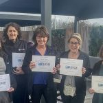 Five smiling women stand outdoors, each holding a certificate that reads