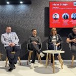 Five panelists are seated on a brightly lit stage in front of a large screen displaying the event title and speaker information. The panel, consisting of four men and one woman, appears to be engaged in a discussion about entrepreneurship and leadership within their industry.